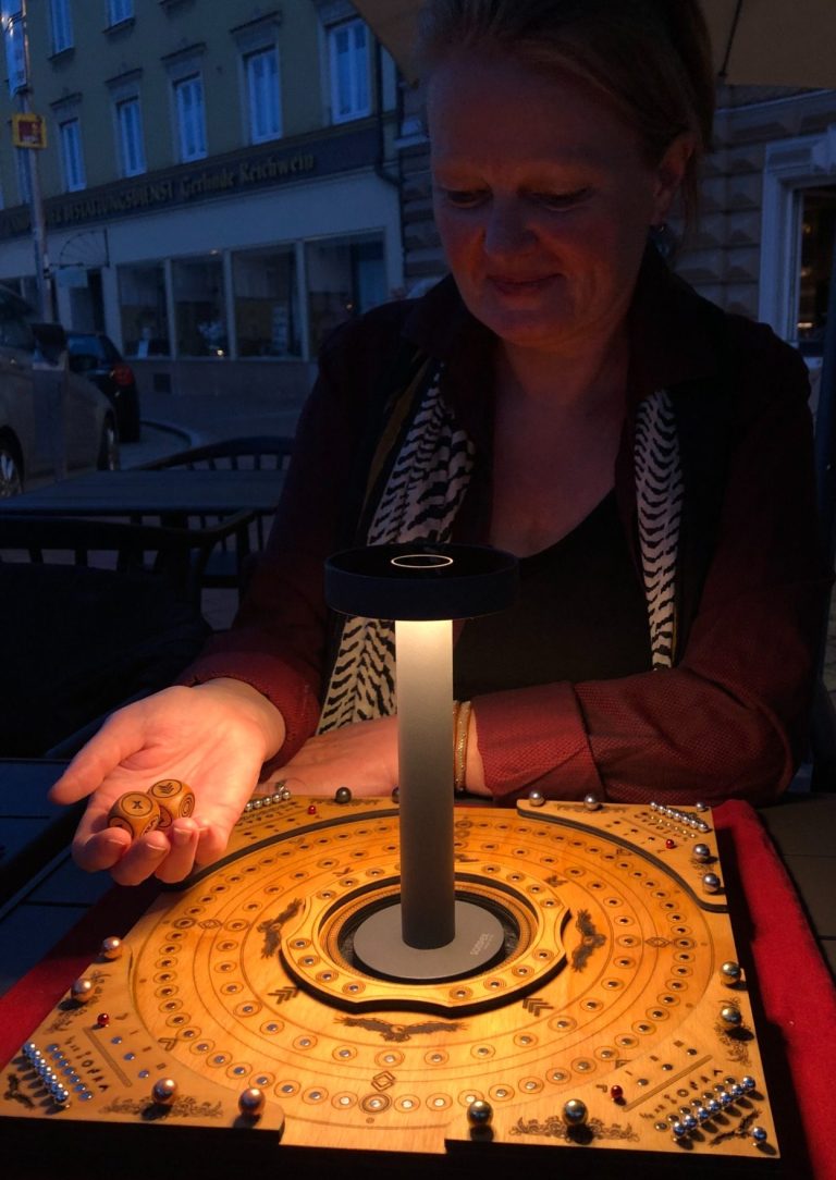 The Inner Circle mit Beleuchtung - Adler Holzhandwerk Eine Dame würfelt zwei Würfel vor dem Brettspiel The Inner Circle, welches auf einem Tisch platziert ist